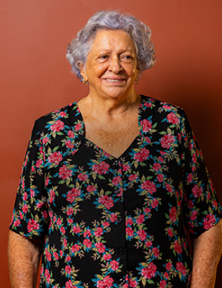 Dona Lourdes sorrindo, de blusa florida, diante de uma parede terracota