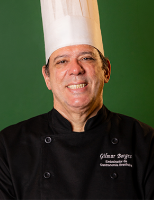 Gilmar Borges sorrindo, de chapéu de chef e dólmã preta, diante de uma parede verde
